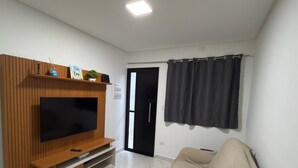Uma sala de estar com televisão de tela plana, unidade de entretenimento de madeira, um sofá bege, uma porta preta e uma janela com cortinas escuras.