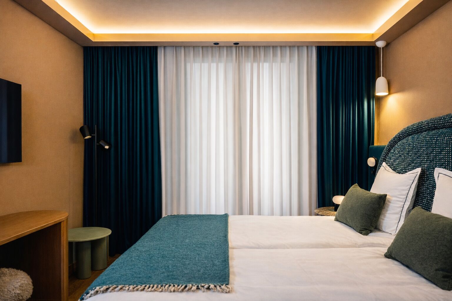 A hotel room with a bed, two bedside tables, a lamp, a TV, and a window with curtains.