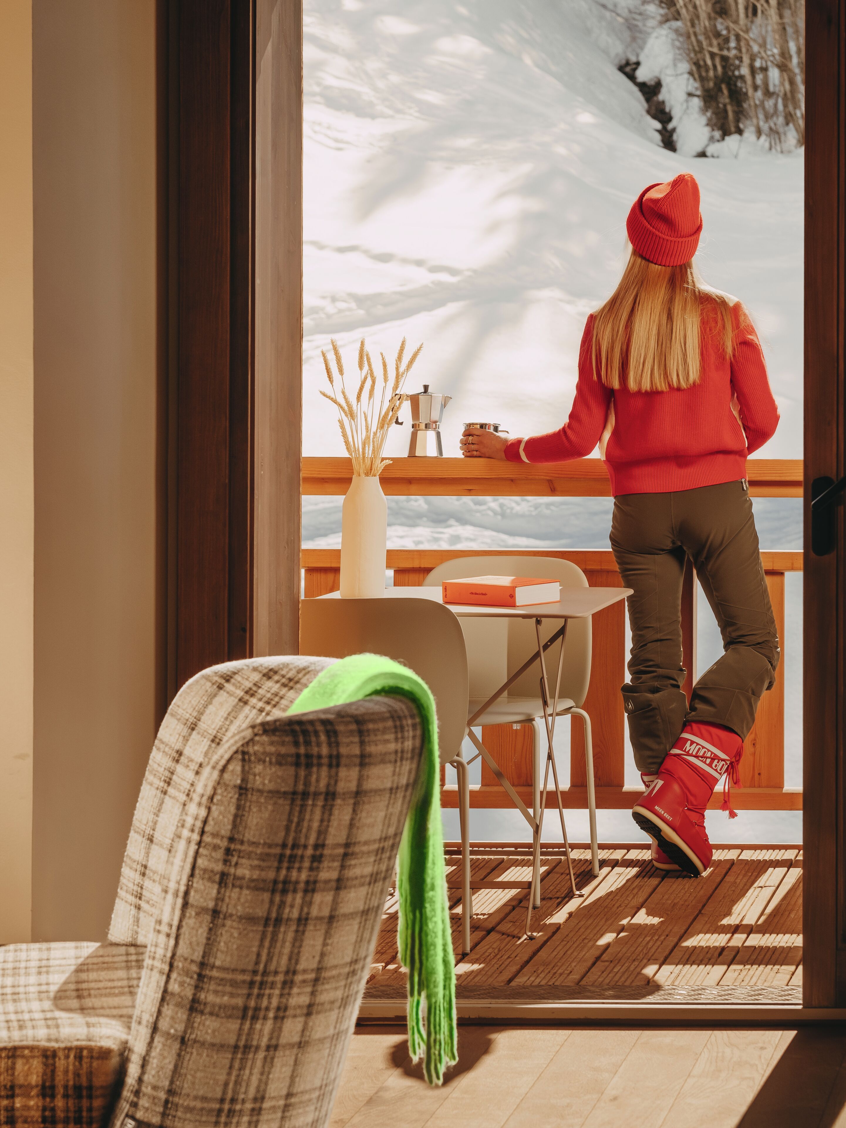 A person standing by a window with a view of a snowy landscape, a coffee maker, and a vase with dried plants on the windowsill.
