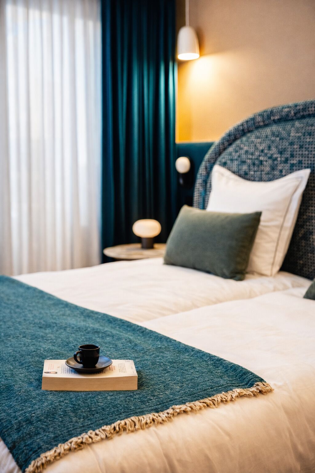 Un lit bien fait, avec une couverture bleue, un oreiller et un livre posé sur lequel se trouve une tasse.