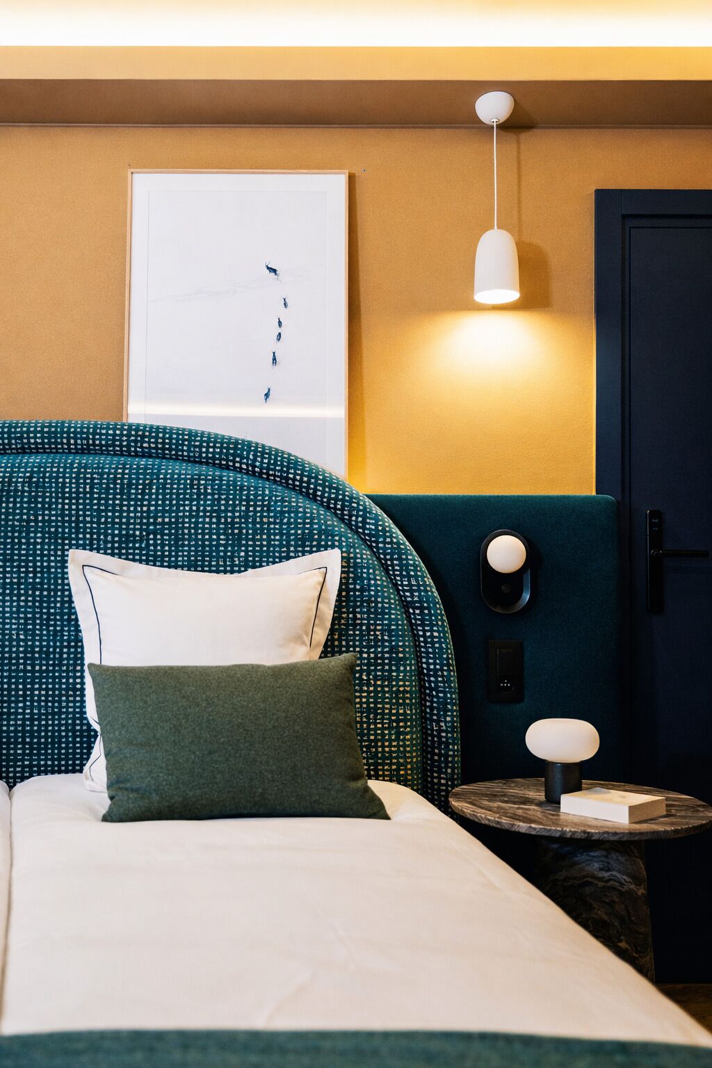 Habitación de hotel ordenada con cama, almohadas, mesita de noche, lámpara, puerta y aplique de pared.