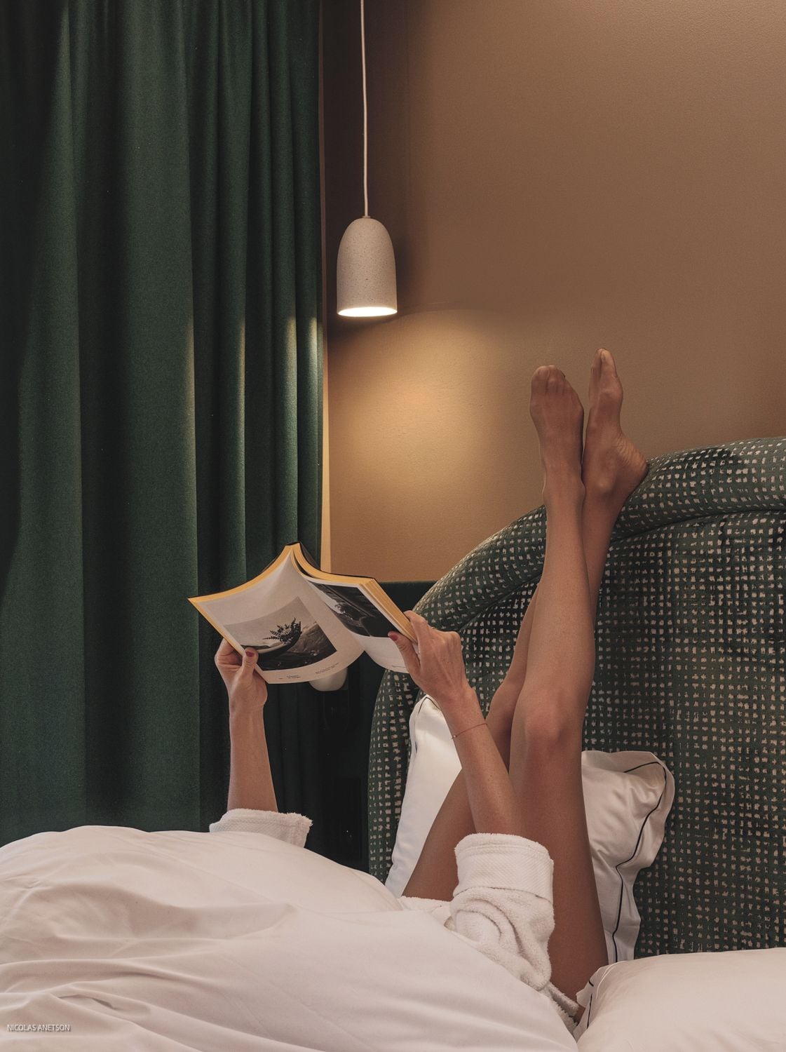 A person lying in bed reading a book with a lamp above.