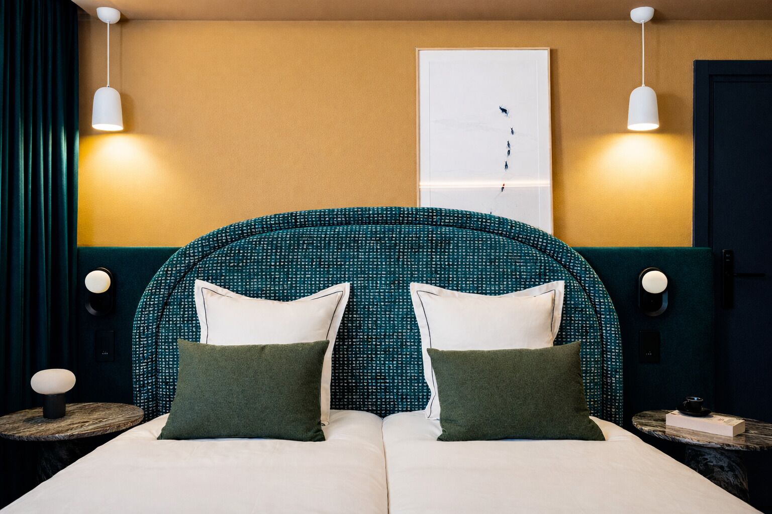 Une chambre d’hôtel avec un grand lit, deux tables de chevet, une lampe fixée au mur et une porte.
