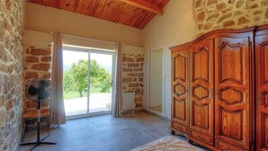Ein Zimmer mit Steinwänden, einer Holzbalkendecke, einem großen Holzschrank, einem Ventilator und einer Glastür, die auf einen Balkon führt.