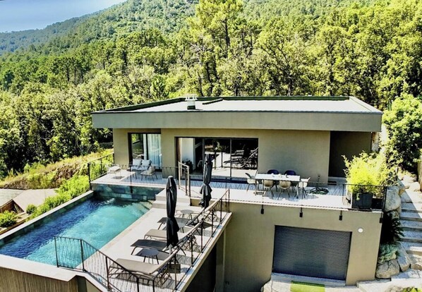 Ein modernes Haus mit Swimmingpool, Sitzgelegenheiten im Freien und Blick auf einen bewaldeten Hang.