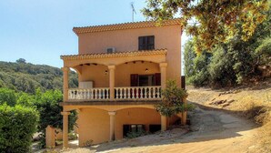 Edifici de dues plantes amb un balcó, envoltat de vegetació i turons.