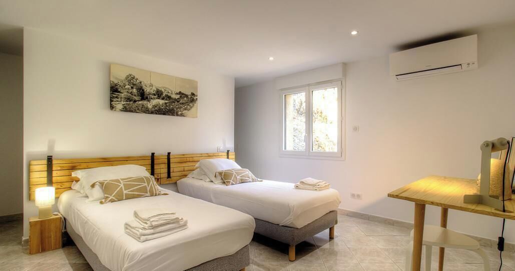 A bedroom with two beds, a bedside table, a lamp, a window, and an air conditioning unit.