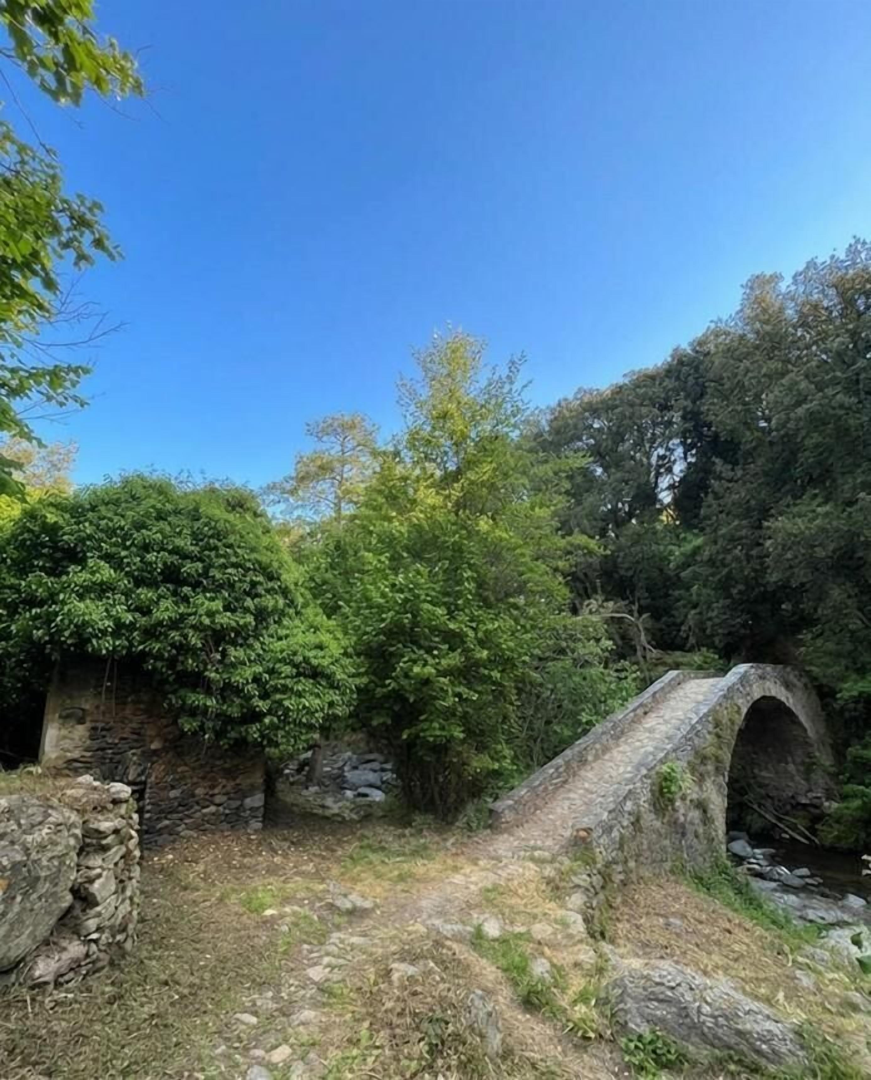 An old stone bridge over a stream, surrounded by dense greenery and a stone wall.
