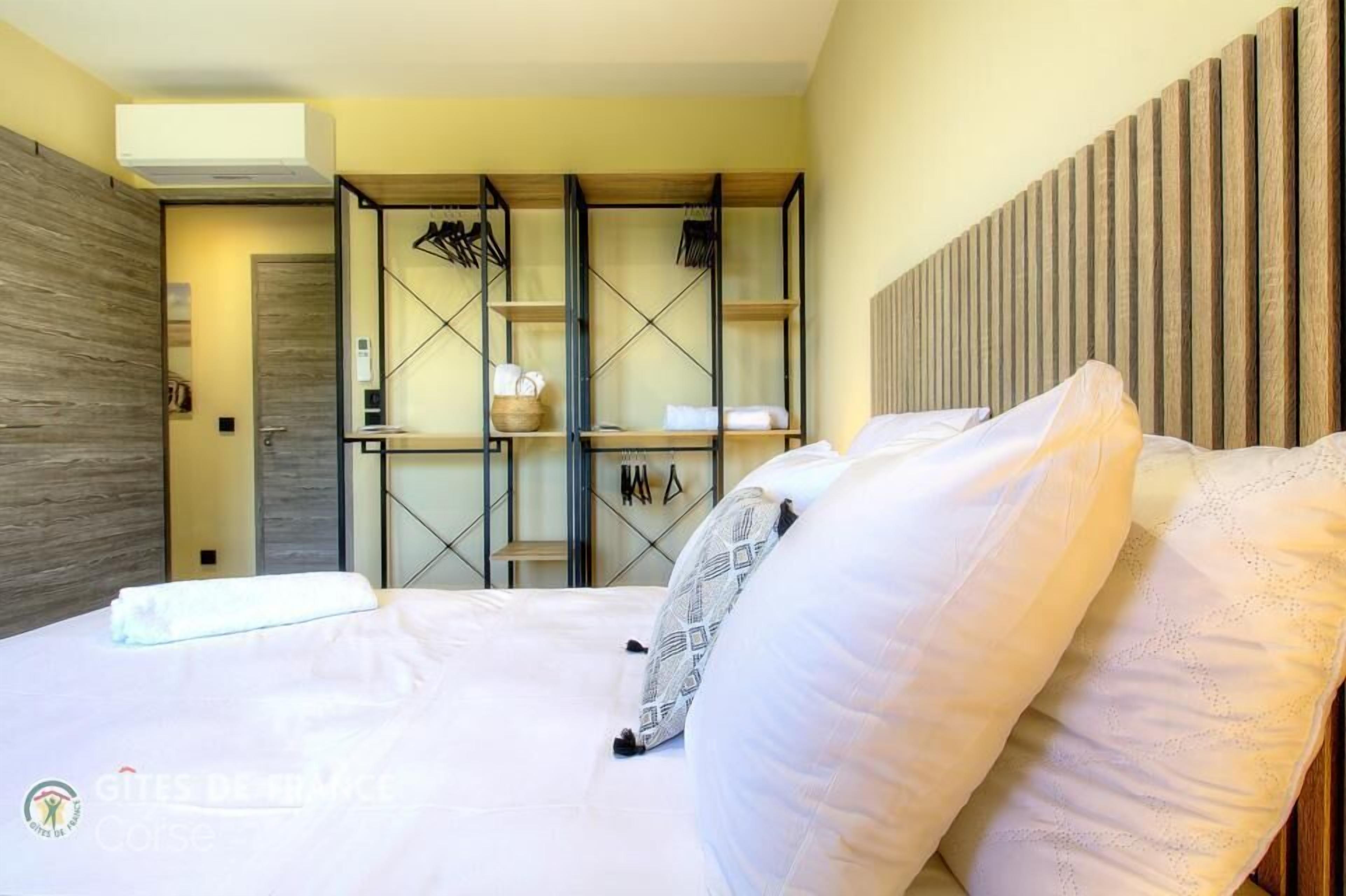 A hotel room with a bed, a wardrobe with a mirror, and a shelf with towels and toiletries.