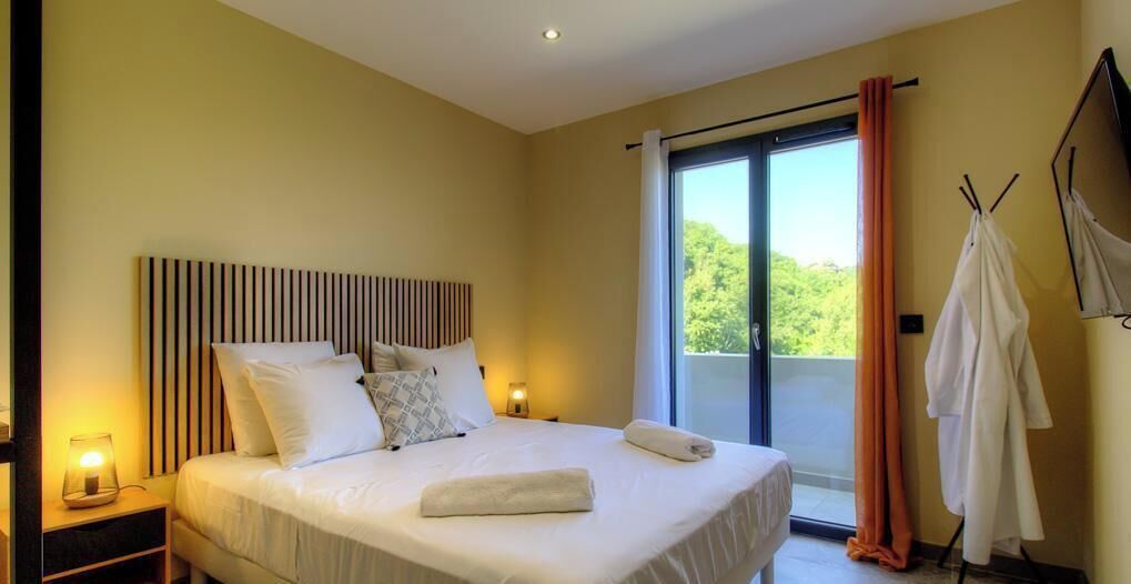 A bedroom with a large bed, a nightstand, a window with curtains, and a view of greenery outside.
