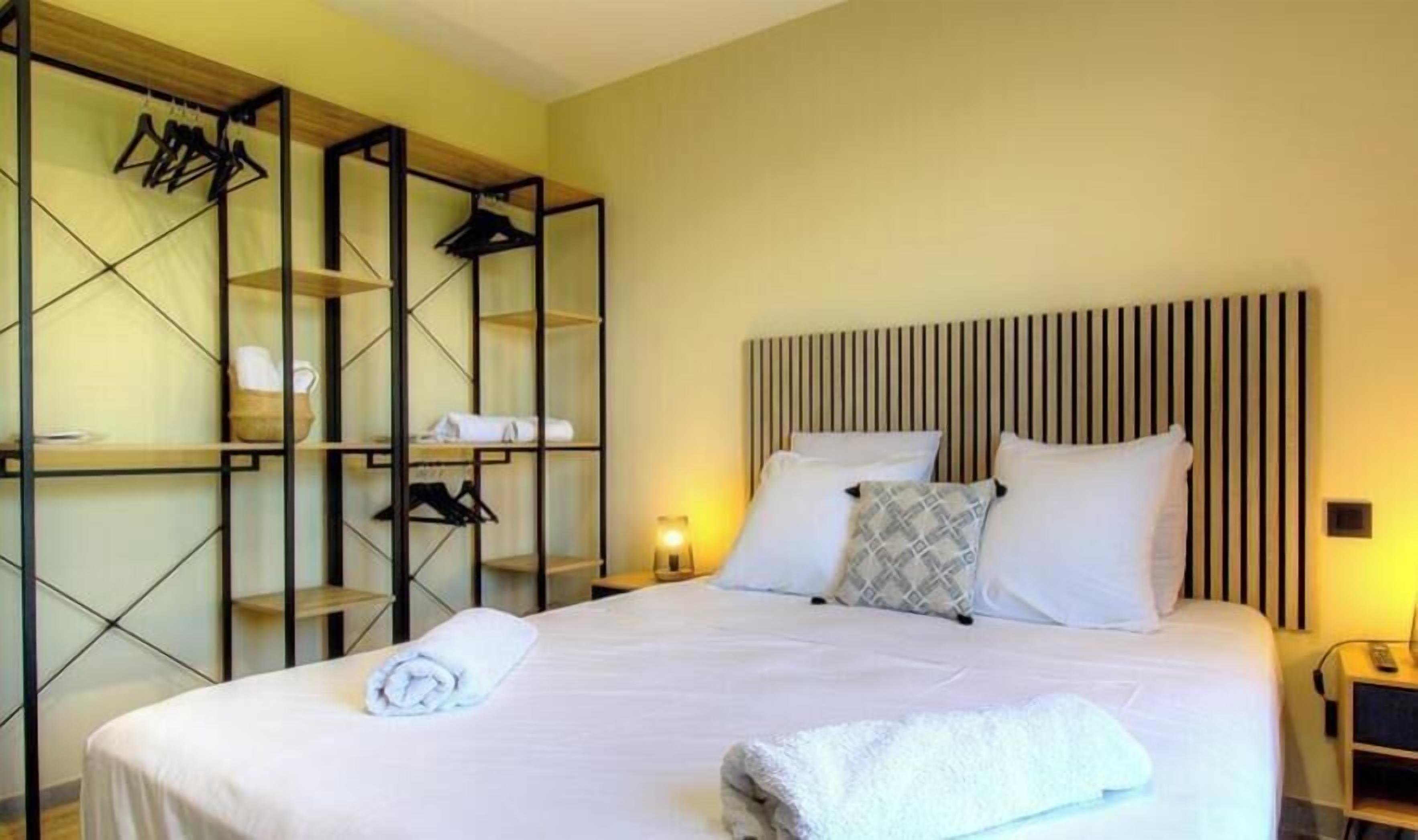 Quarto de hotel com cama, guarda-roupa cheio de roupas penduradas e mesa de cabeceira com abajur.