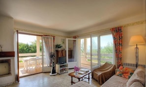 Ein Wohnzimmer mit Sofa, Couchtisch, Fernseher und Ventilator. Eine Glastür führt auf einen Außenbereich mit Tisch und Stühlen.