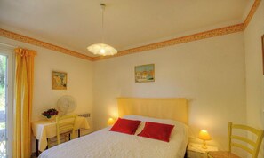 Ein Schlafzimmer mit einem Bett, zwei Nachttischlampen, einem Ventilator, einem Tisch mit Stuhl und zwei Bildern an der Wand.
