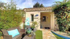 Una casa con techo de tejas, una escultura de caballito de mar azul, un sofá de mimbre con cojín azul y un camino de piedra que lleva a la entrada.