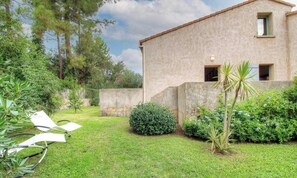 Una casa de piedra con un ventanal pequeño y un jardín con césped, una palmera y una silla.