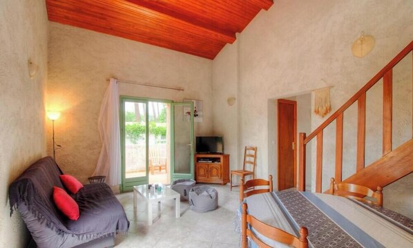 Una habitación con techo de madera, un sofá con cojines rojos, un televisor, una mesa de comedor con sillas y una escalera.