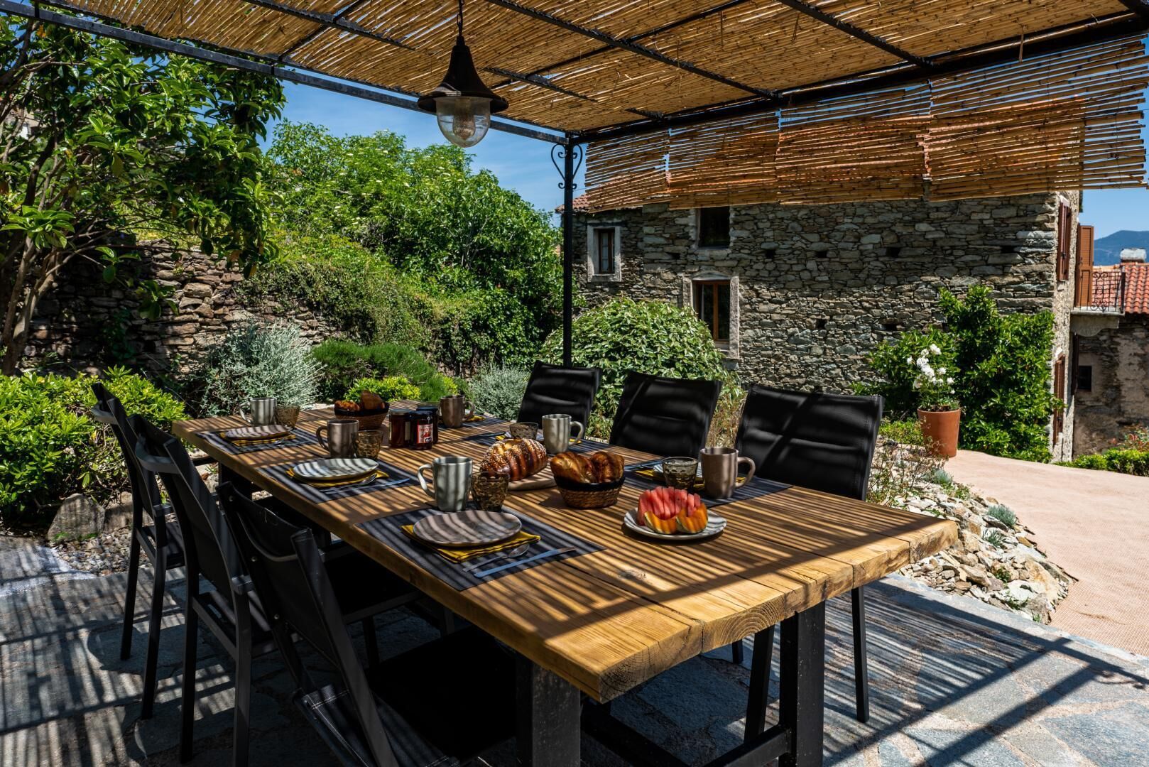 Un espace de restauration en plein air, avec un mur en pierre en arrière-plan, une table en bois dressée avec des assiettes, des tasses et de la nourriture, et un luminaire suspendu.