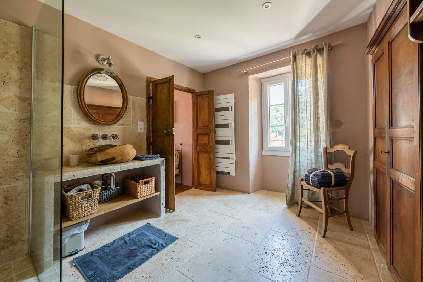 A bathroom with a glass shower enclosure, a wooden vanity with a mirror, a towel rack, and a chair.