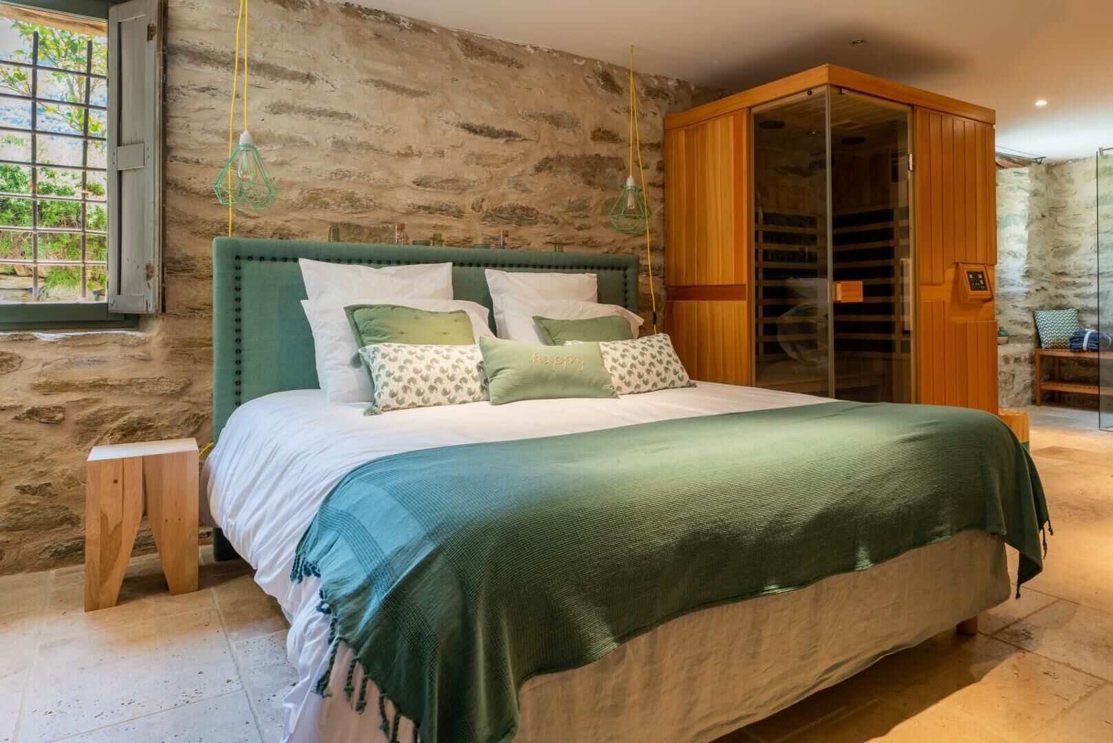 Una camera da letto con un letto, un comodino in legno, una testiera imbottita verde e un muro in pietra.