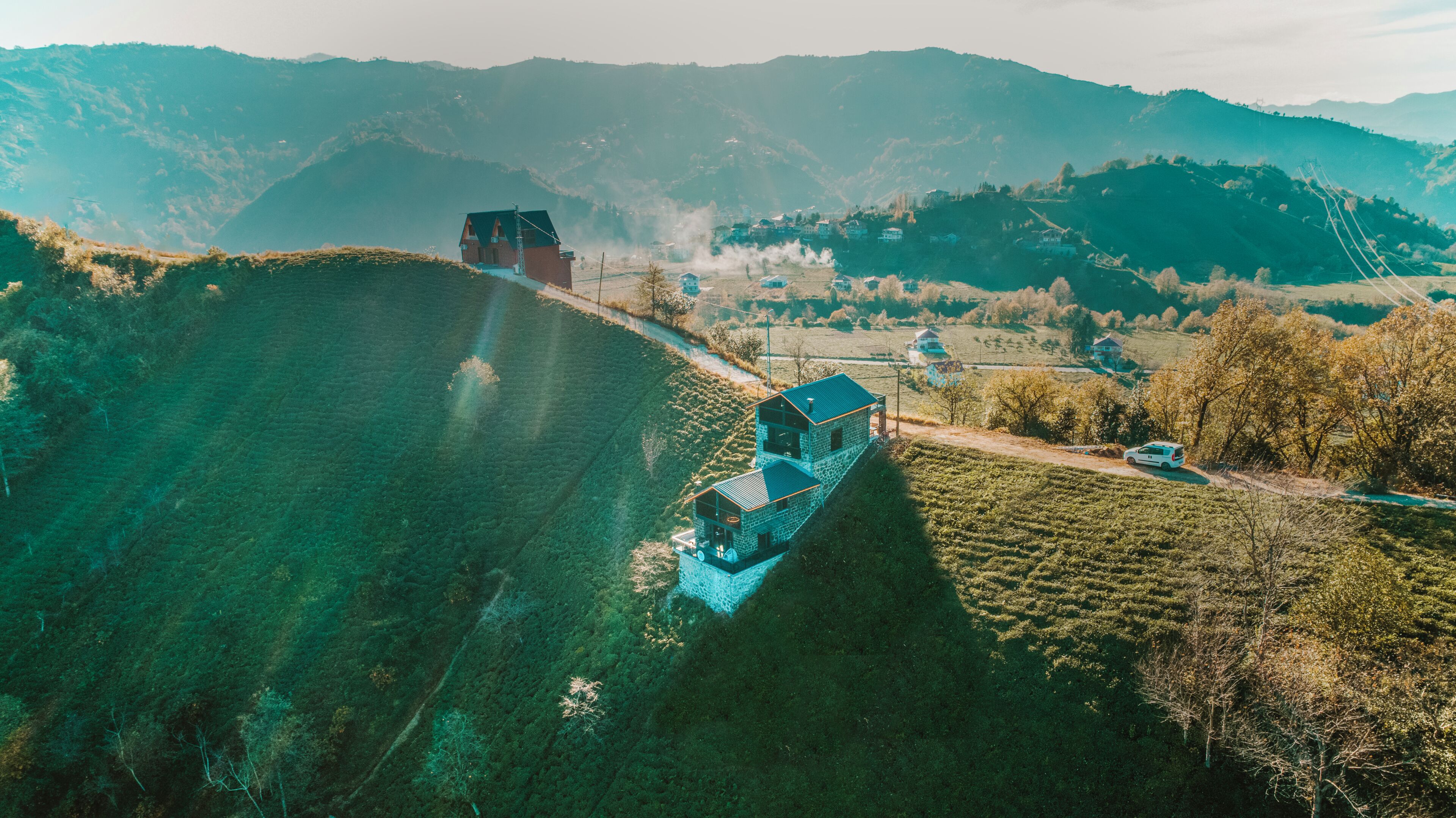 A large, multi-story stone building with a flat roof, situated on a hillside with a view of surrounding hills and a small stream.