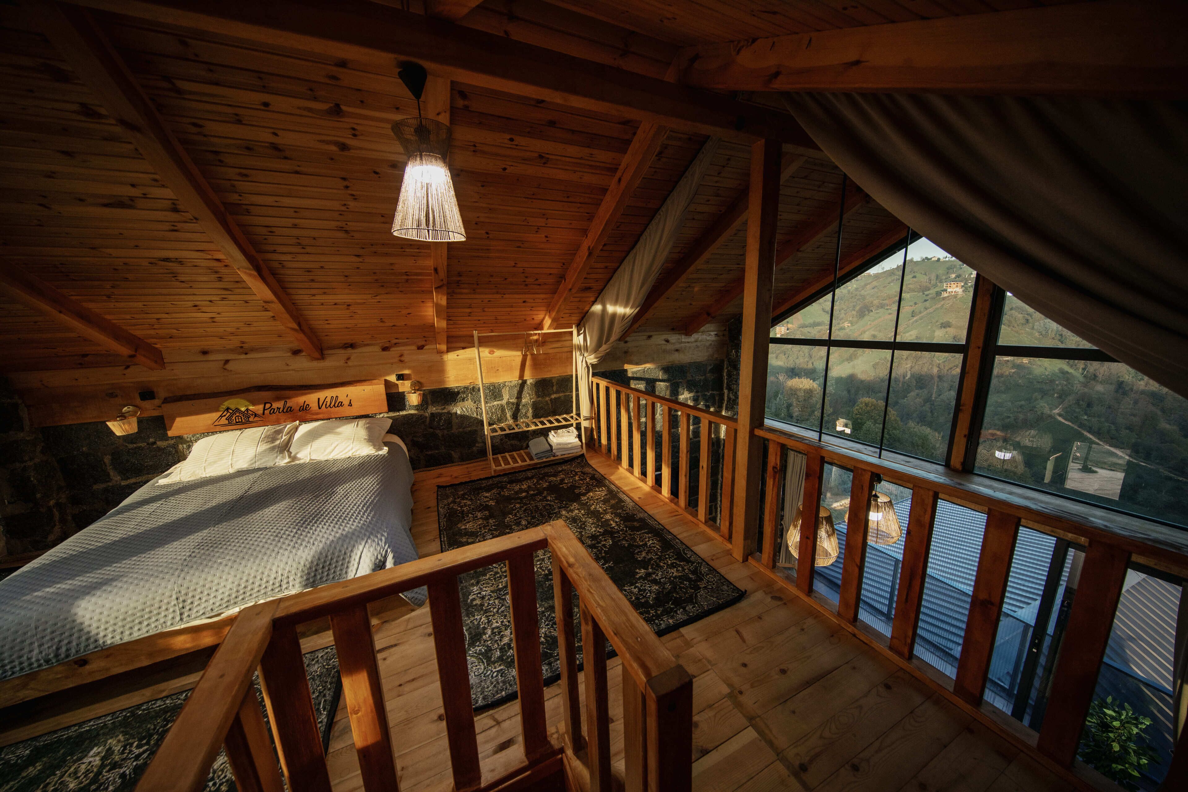 A wooden cabin interior with a bed, a stone fireplace, a view through large windows, and a wooden staircase.