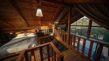 A wooden cabin interior with a bed, a stone fireplace, a view through large windows, and a wooden staircase.