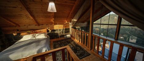 A wooden cabin interior with a bed, a stone fireplace, a view through large windows, and a wooden staircase.