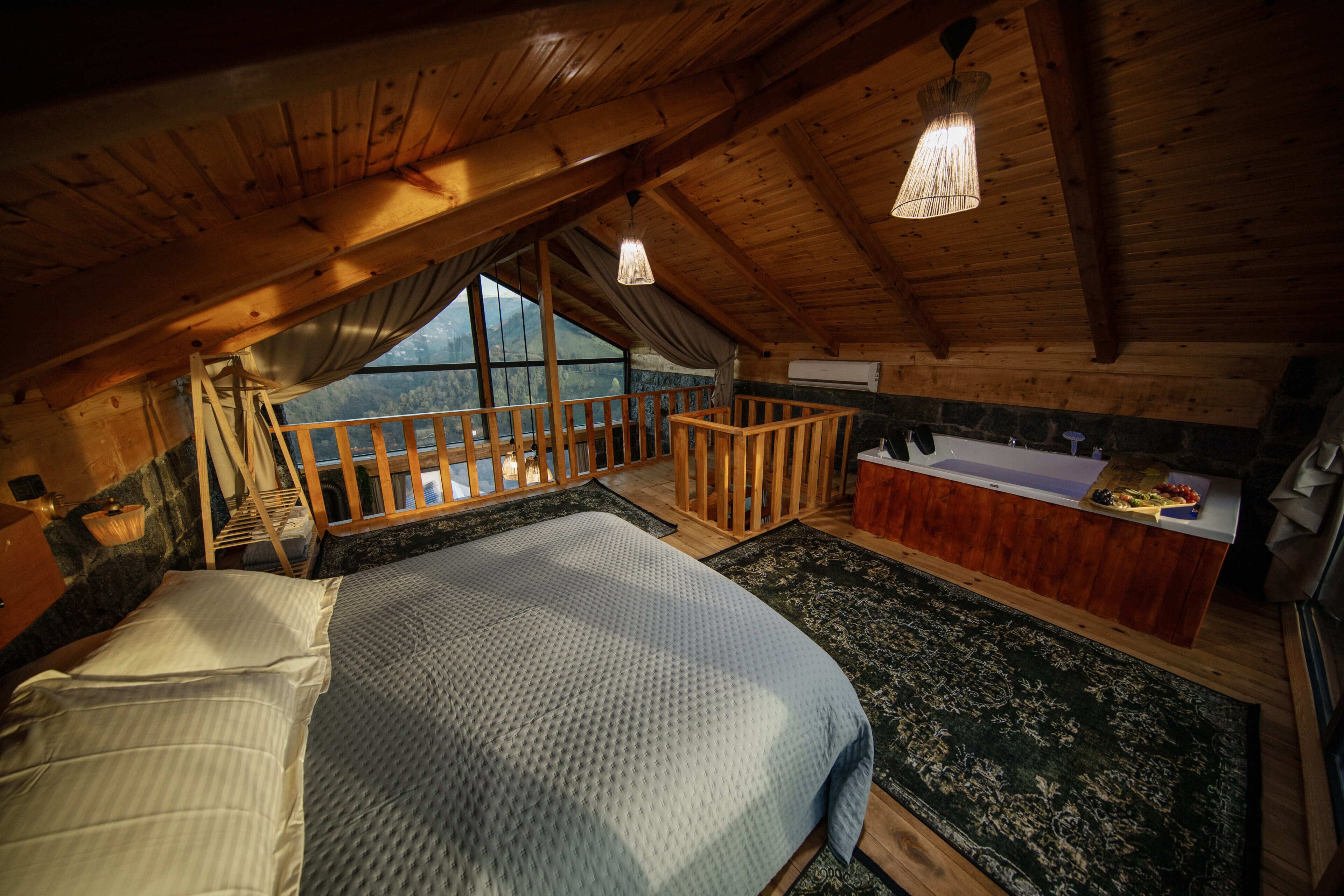 A bedroom with a large bed, a wooden balcony, a bathroom with a bathtub, and a view of the mountains.