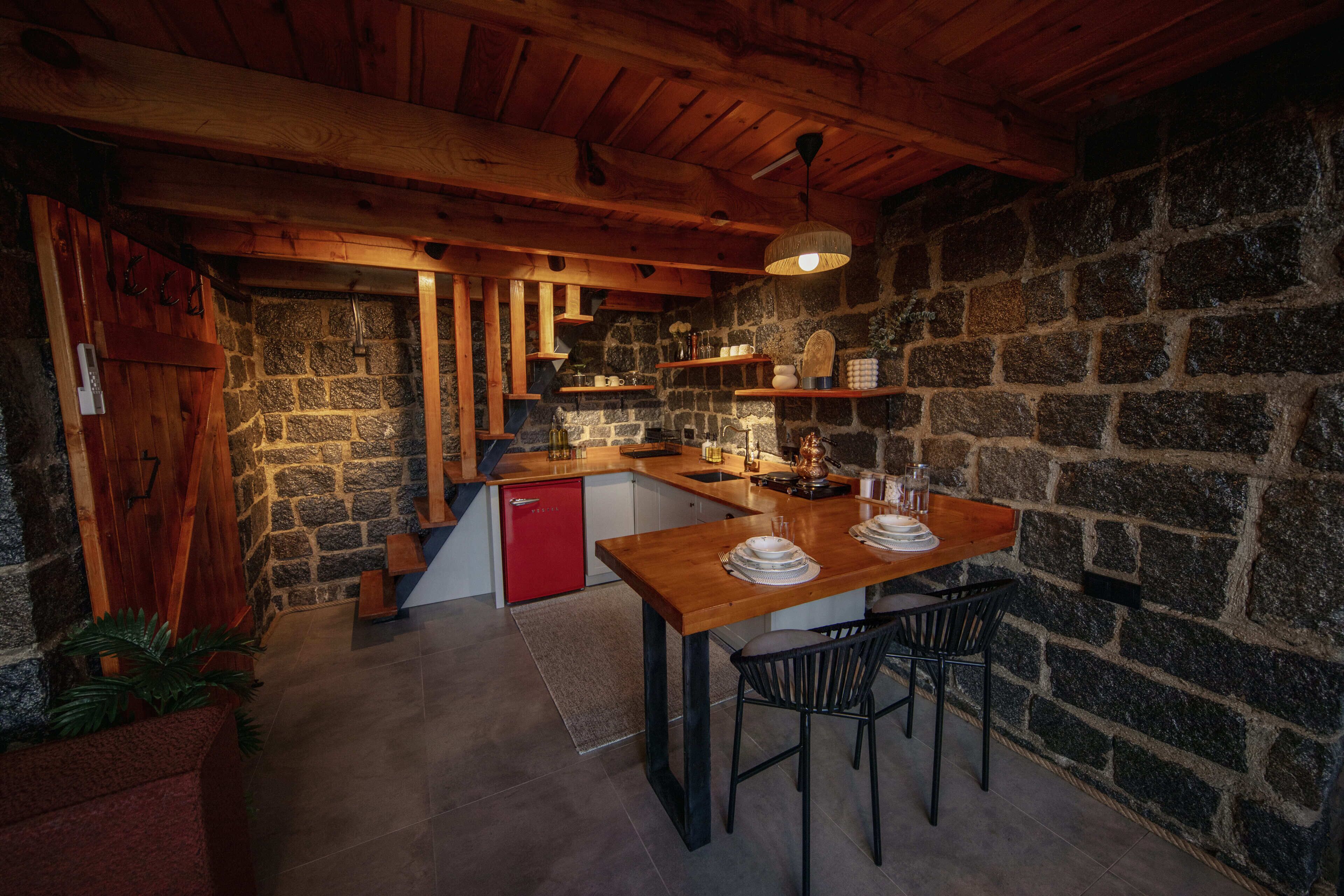 A rustic kitchen with stone walls, wooden beams, a red refrigerator, and a wooden dining table with chairs.