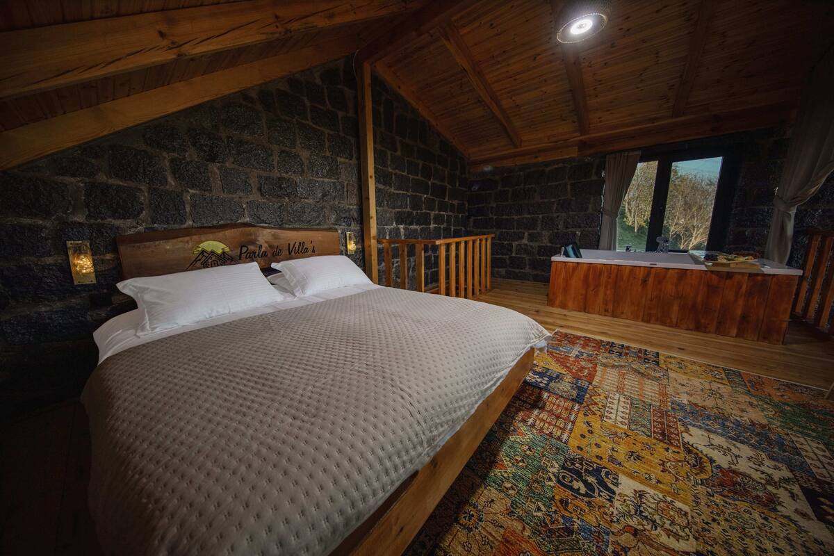 Un dormitorio con pared de piedra, techo de madera, cama, banca y vista al exterior.