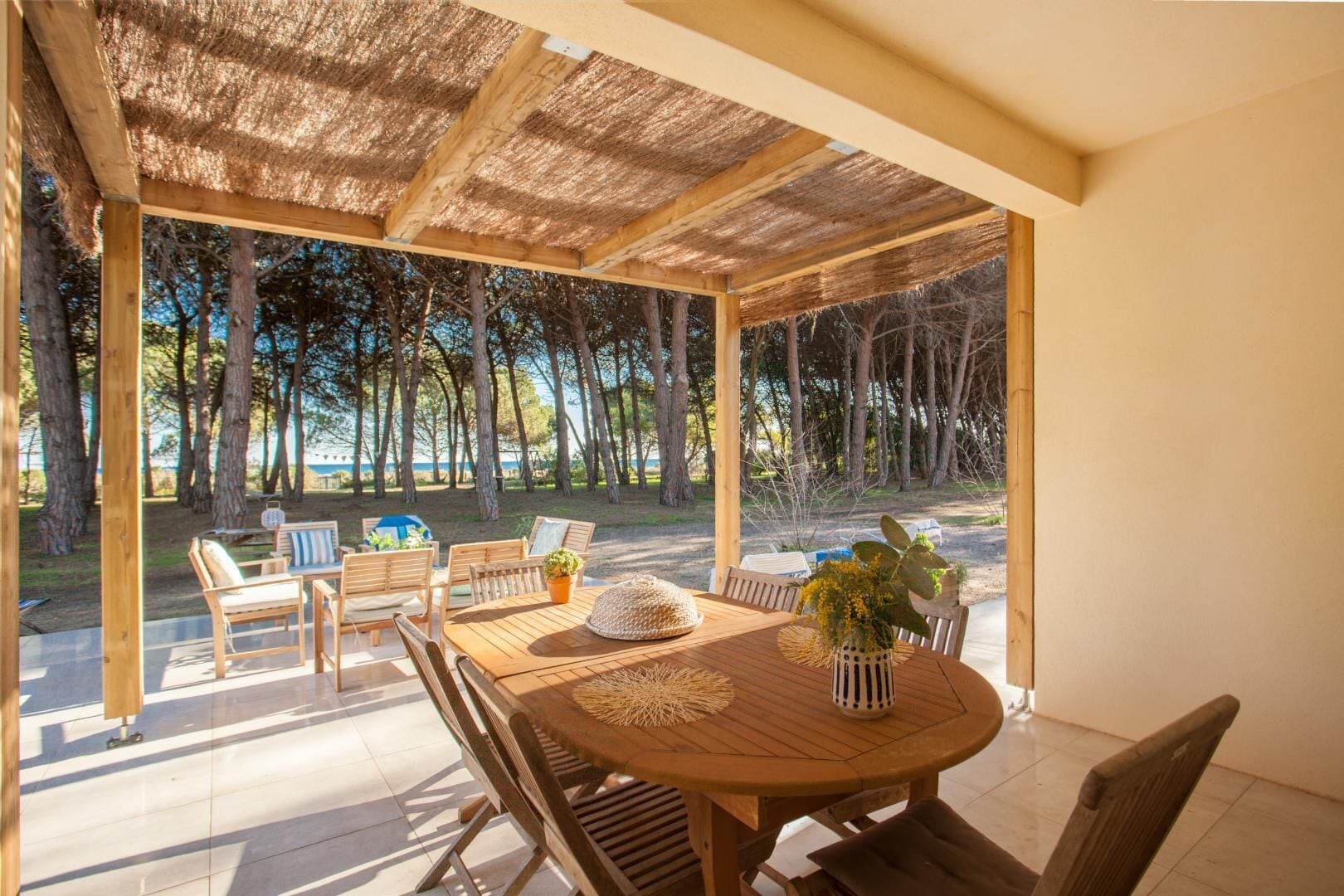 Un espace de restauration en plein air avec une table et des chaises en bois, entouré d’une terrasse couverte d’où l’on peut admirer une forêt et un plan d’eau.
