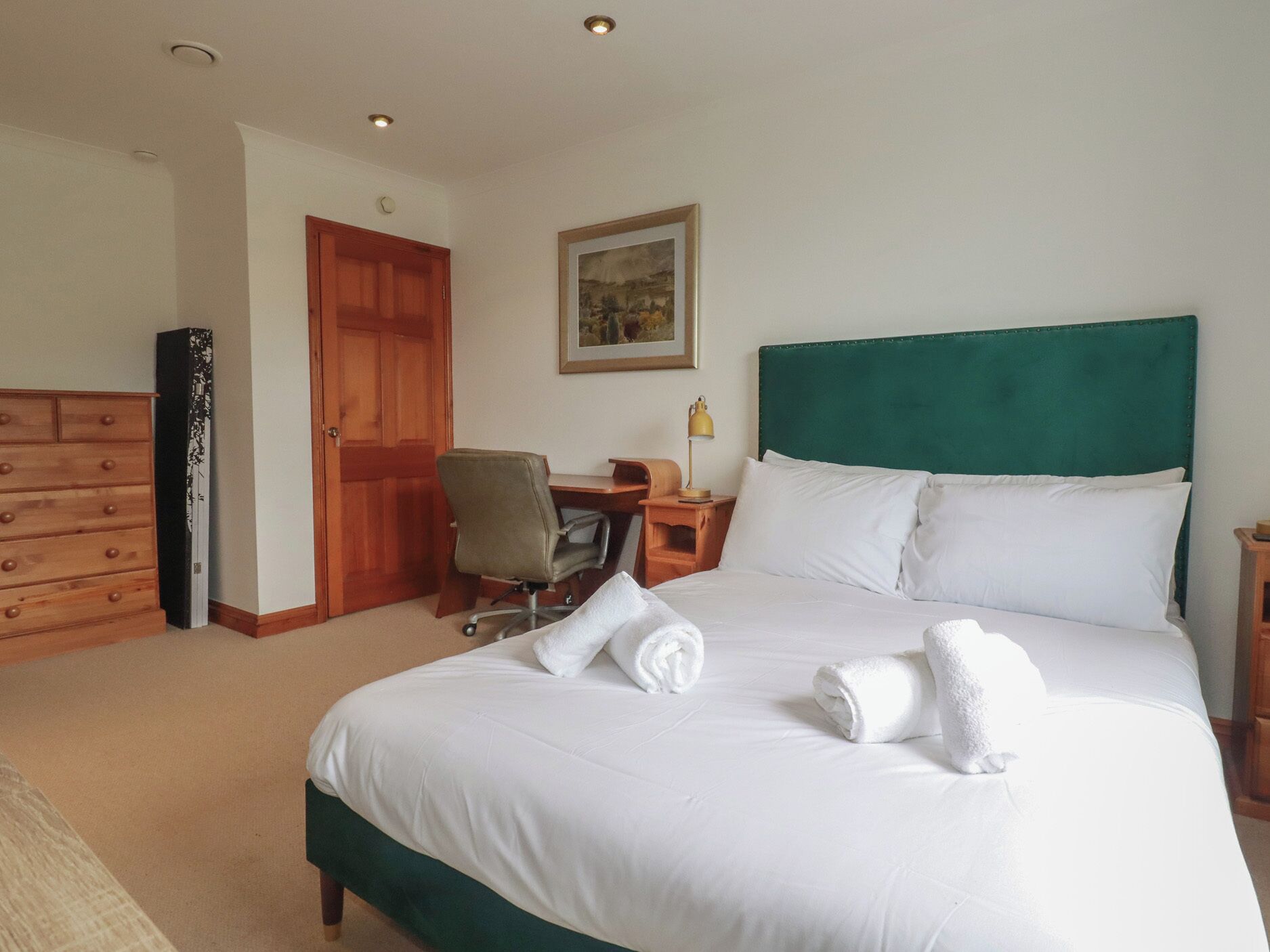 Quarto de hotel com cama, escrivaninha, cadeira, cômoda e porta.