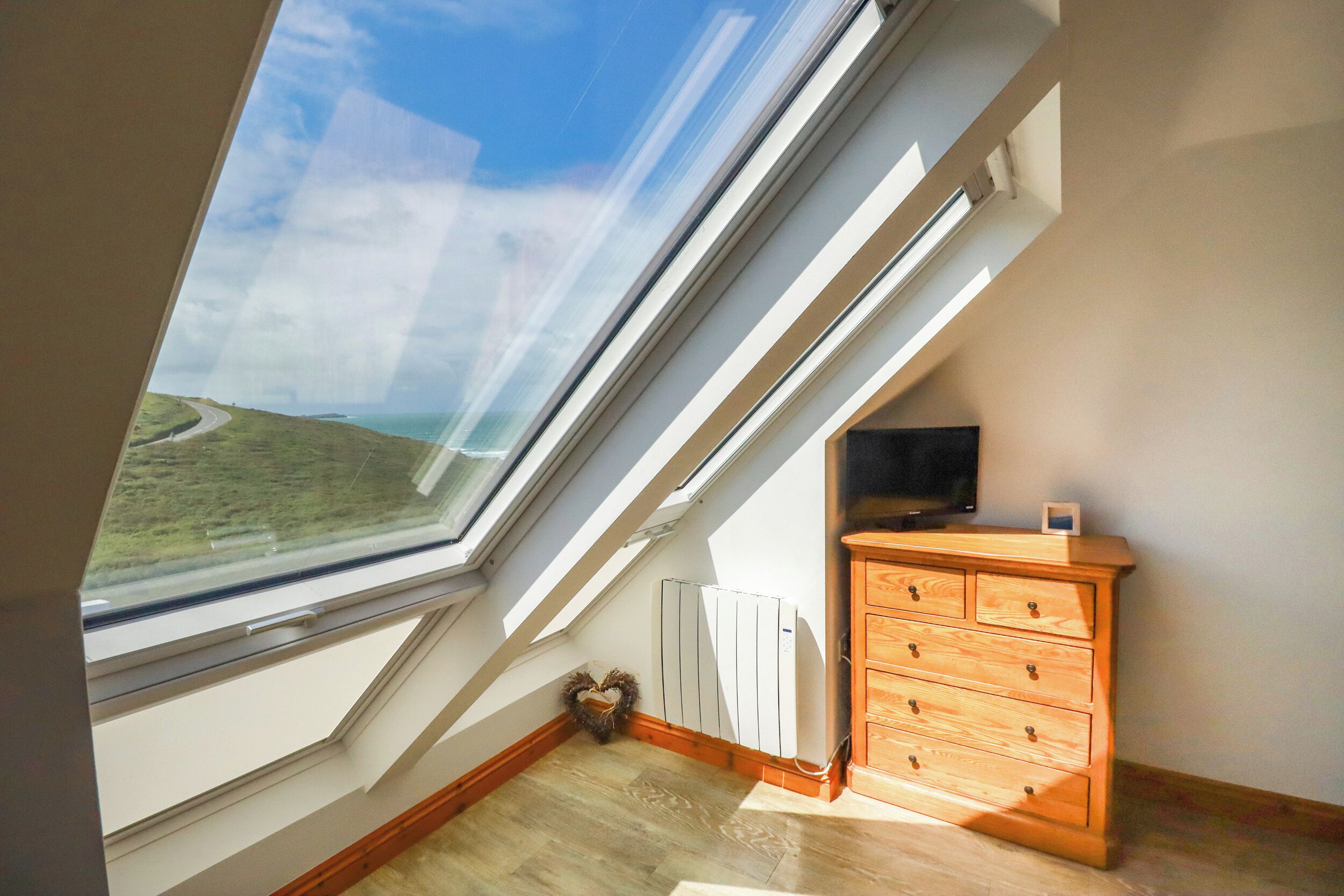 A room with a sloped ceiling, a wooden dresser, a radiator, and a view of a coastal landscape through a skylight.