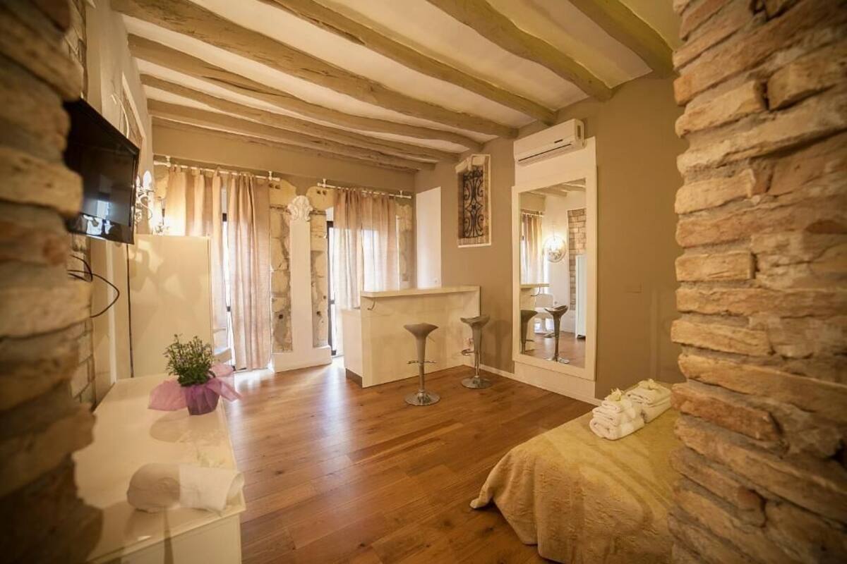 Una habitación con suelo de madera, una cama, un televisor fijado en una pared de piedra, una mesita con una planta y un espejo en la pared.