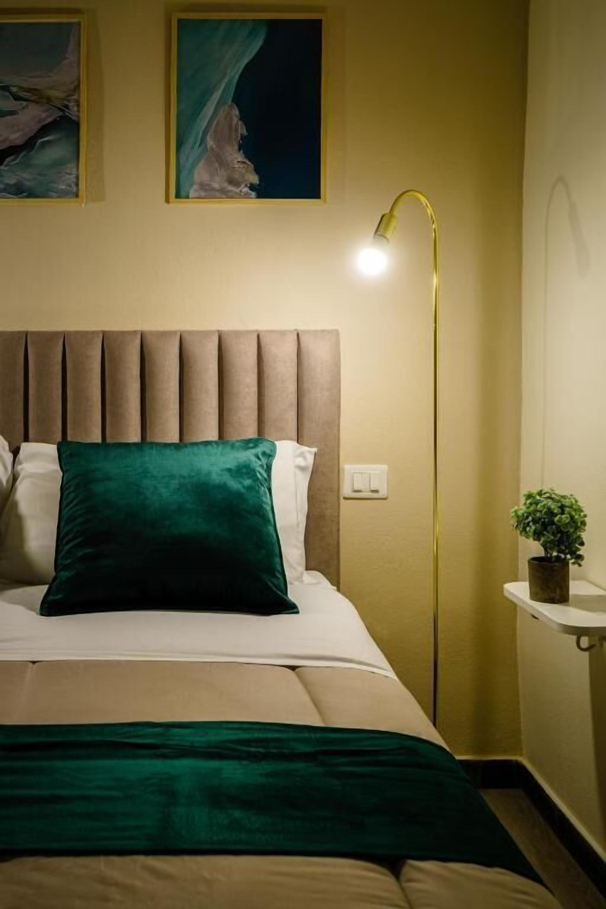 Ein Bett mit Kopfteil, einem grünen Kopfkissen, einer Nachttischlampe und einer kleinen Pflanze auf einem Nachttisch.
