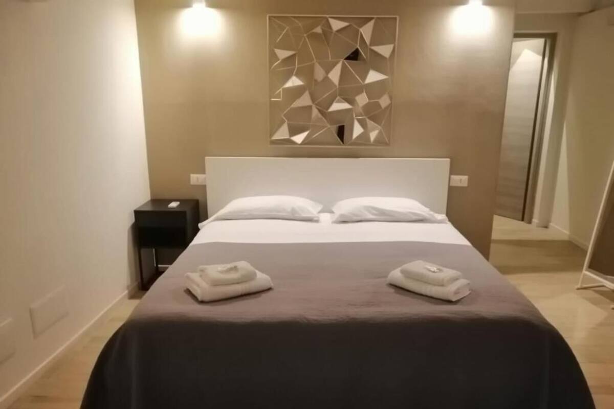 Una habitación de hotel con cama, dos mesitas de noche, un aplique geométrico fijado a la pared y una puerta que da a otra estancia.