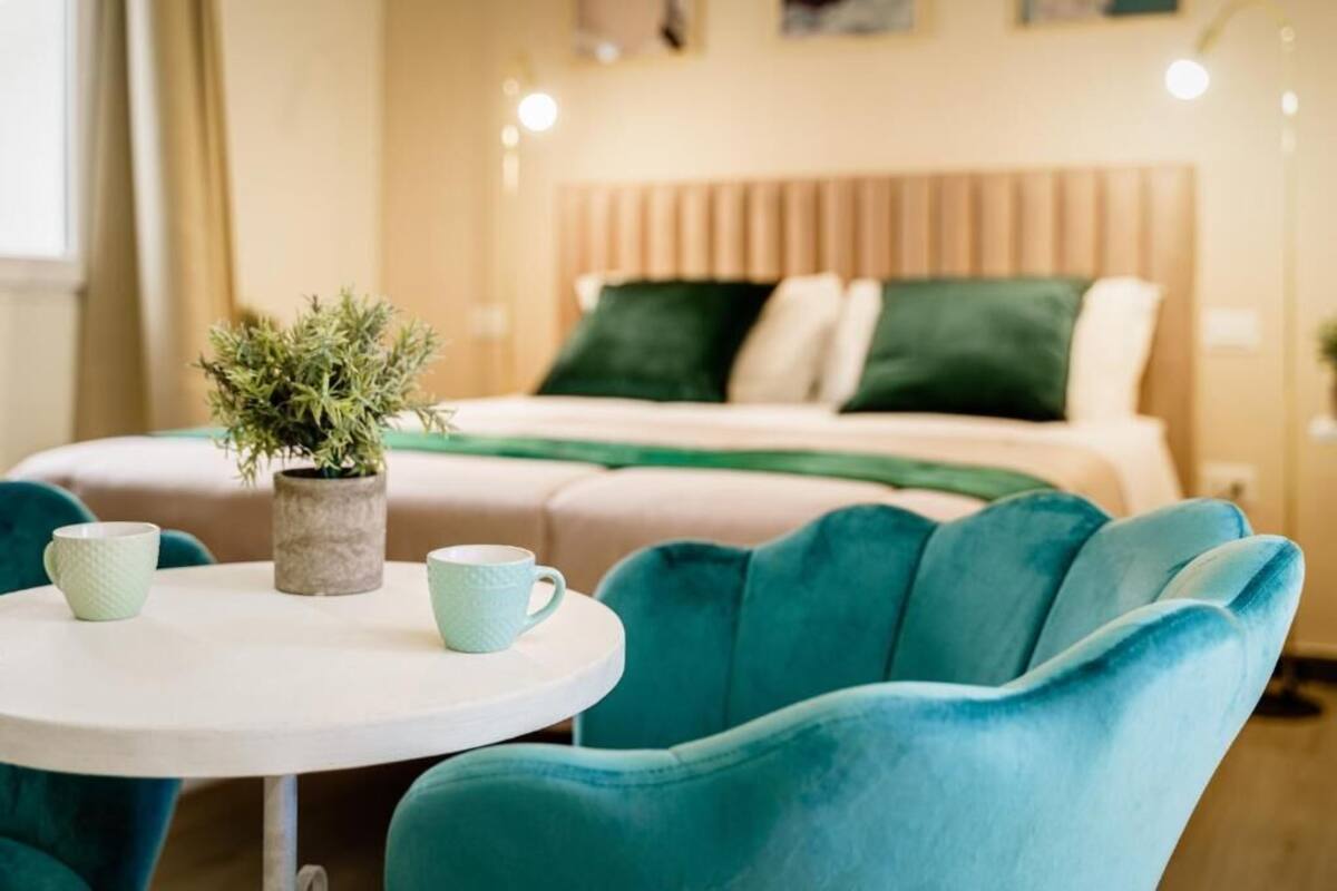 Una cama con ropa de cama verde y blanca, una planta pequeña en maceta y sillas color turquesa alrededor de una mesa blanca.