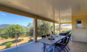 Ein überdachter Balkon im Freien mit Tisch und Stühlen, der einen Blick auf eine Landschaft mit Bäumen und sanften Hügeln bietet.