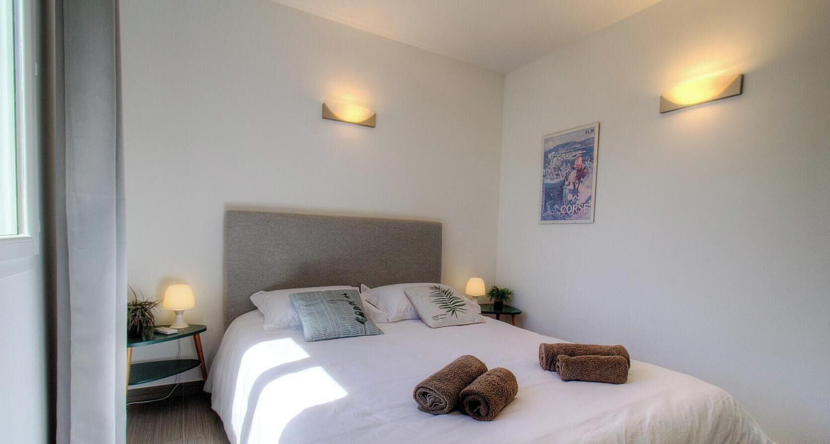 A bedroom with a bed, pillows, a headboard, bedside tables with lamps, a poster on the wall, and rolled towels on the bed.