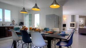 Une cuisine moderne avec un coin repas, agrémentée de luminaires suspendus, d’un grand meuble gris et offrant une vue sur l’extérieur.