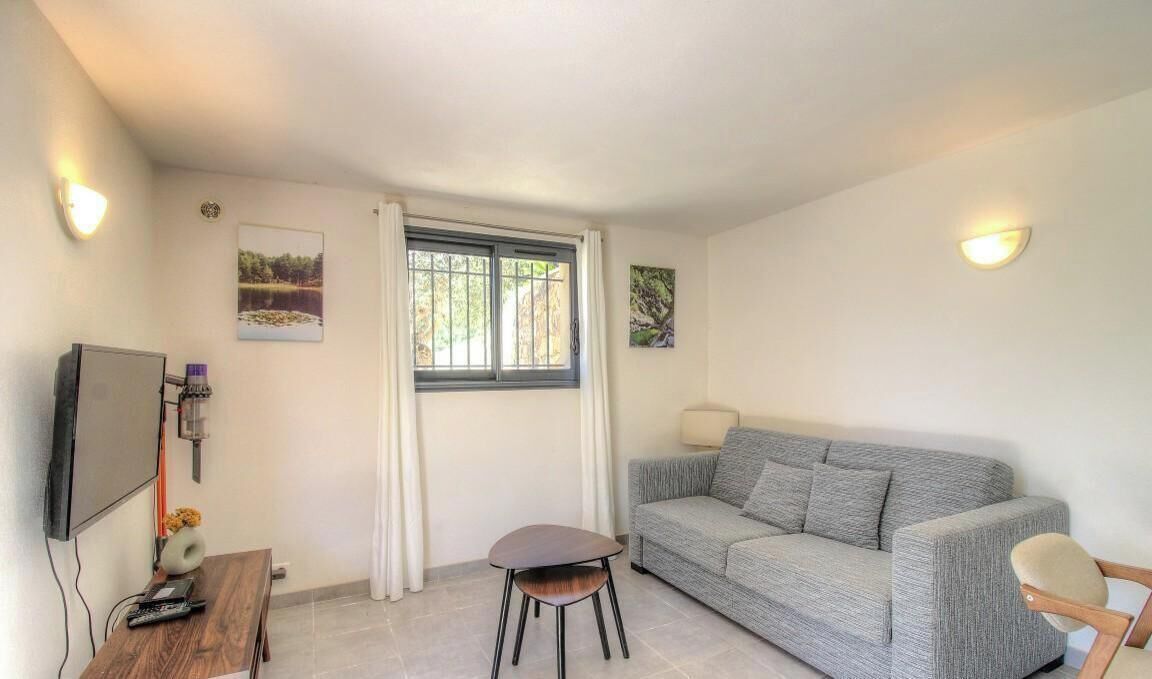 A room with a grey sofa, a small wooden table, a TV mounted on the wall, and a window with curtains.