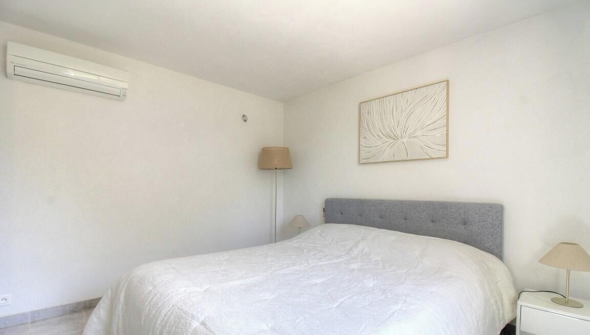 A bedroom with a bed, a nightstand, a lamp, a wall-mounted air conditioner, and a wall art piece.