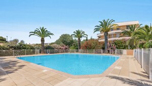 Ein rechteckiger Pool mit Fliesenveranda, umgeben von Palmen und blühenden Sträuchern, mit einem mehrstöckigen Gebäude im Hintergrund.