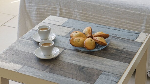 Ein Bett mit weißer Bettdecke und einem blau gestreiften Kopfkissen, ein kleiner Holztisch mit einer Auswahl an Gebäck und zwei Tassen Kaffee.
