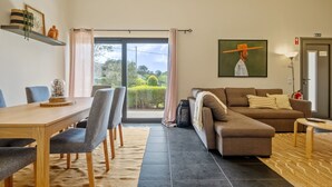 A modern living room with a sofa, a dining table with chairs, a large window with curtains, and a painting on the wall. - Sweet Orchard Escape - By Wehost (Porto Moniz)