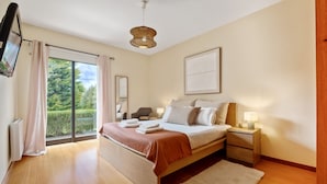 A modern bedroom with a large bed, bedside tables, a flat-screen TV, and a view of greenery through the window. - Sweet Orchard Escape - By Wehost (Porto Moniz)