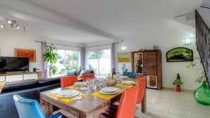 Uma sala de jantar com mesa de madeira posta para quatro pessoas, rodeada por cadeiras laranja, com uma televisão e um sofá ao fundo.