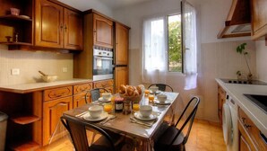 A kitchen with wooden cabinets, a dining table set for breakfast, and a window with curtains. - Casa Margone - CALVI (CALVI)