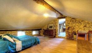 Une chambre mansardée chaleureuse avec un plafond en pente, des poutres en bois, un lit recouvert d’une couverture bleue, une commode et un miroir.