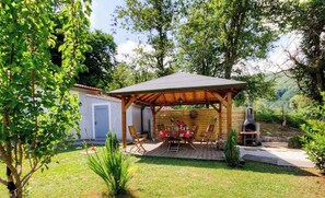 Eine überdachte Terrasse im Freien mit Grill, Holzstühlen und einem kleinen Schuppen, eingebettet in einen üppigen Garten.
